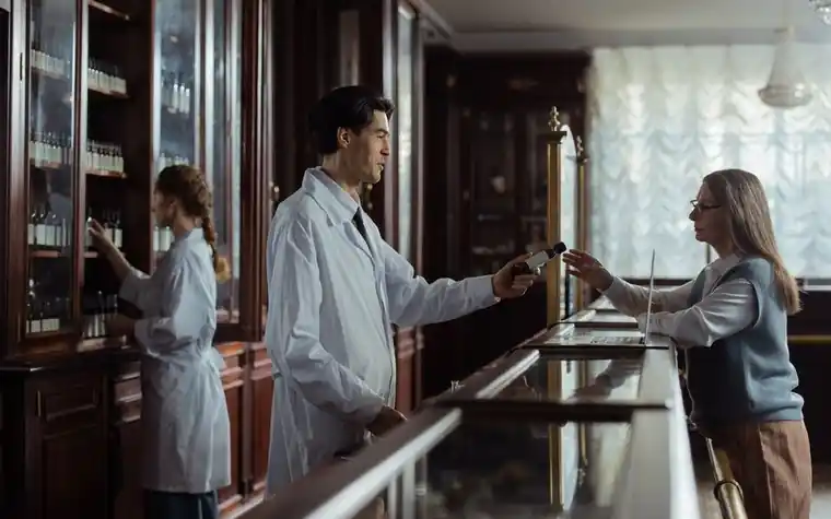 Pharmacist in a white coat handing a medication to a customer across a wooden pharmacy counter