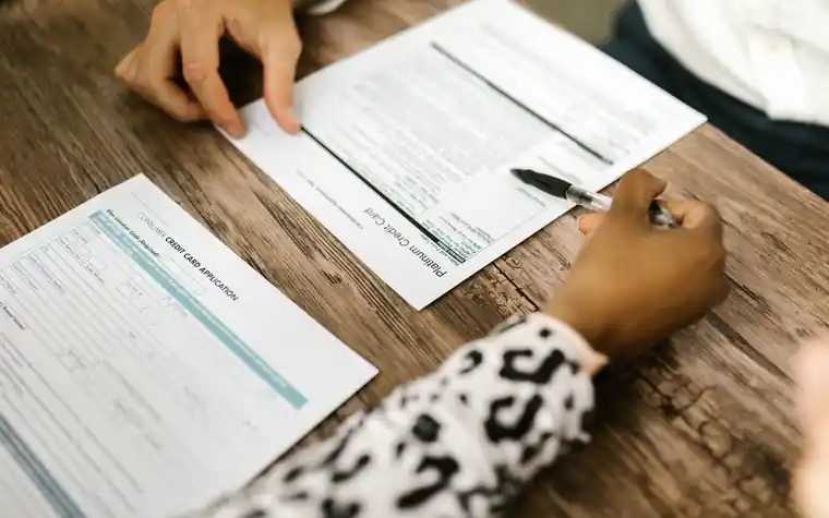 Two people filling out printed forms with pens at a wooden table