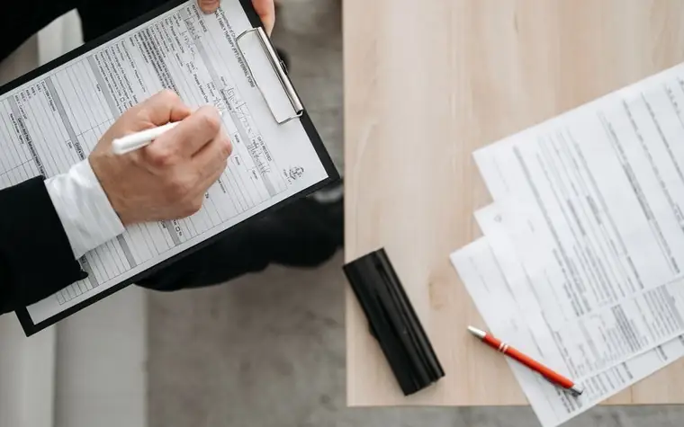 Person filling out a form on a clipboard with more papers on a table nearby