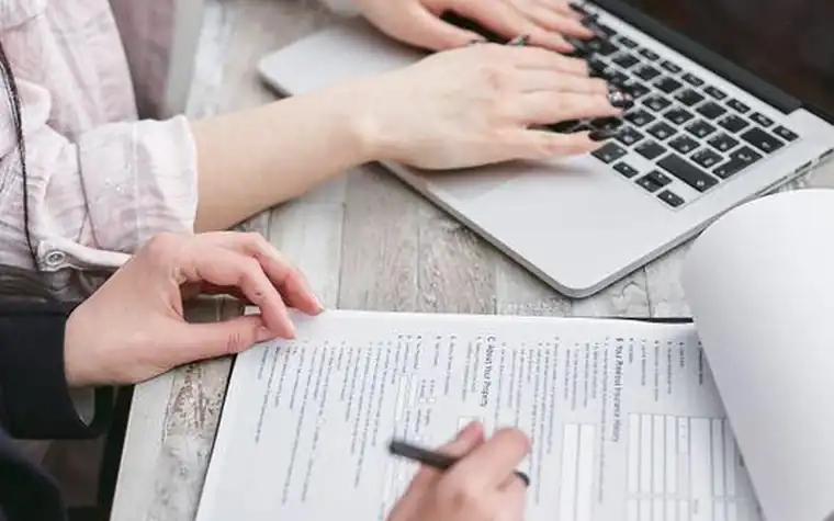 Hands typing on a laptop while another hand fills in a paper form with a pen