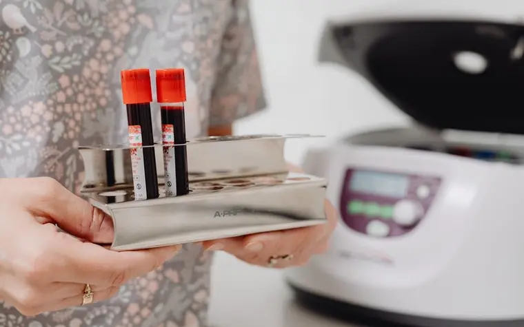 Gloved hands holding a rack of blood sample tubes beside a laboratory centrifuge