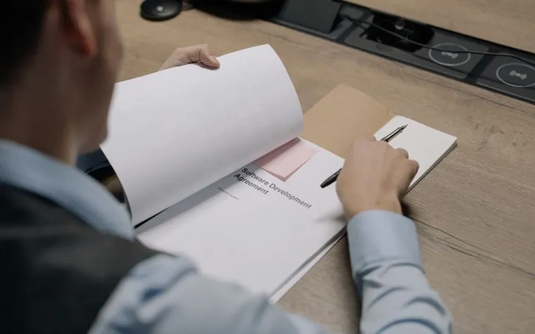 Hand holding a pen over a folder of application paperwork on a desk