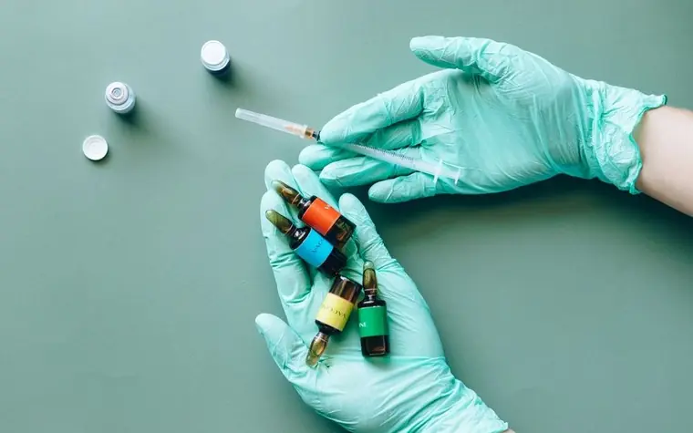 Gloved hands holding small colored medical vials and a syringe on a green background