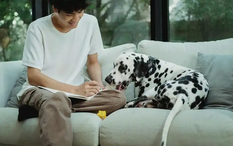 A man writing notes beside a spotted dog on a sofa