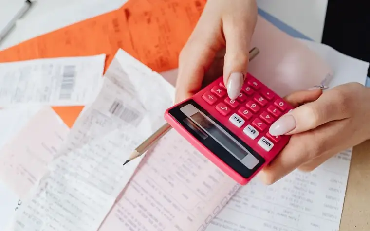 Hands using a pink calculator on a desk covered with receipts to total expenses