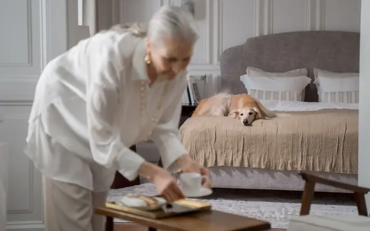 A senior woman setting a tray near her dog resting on a bed