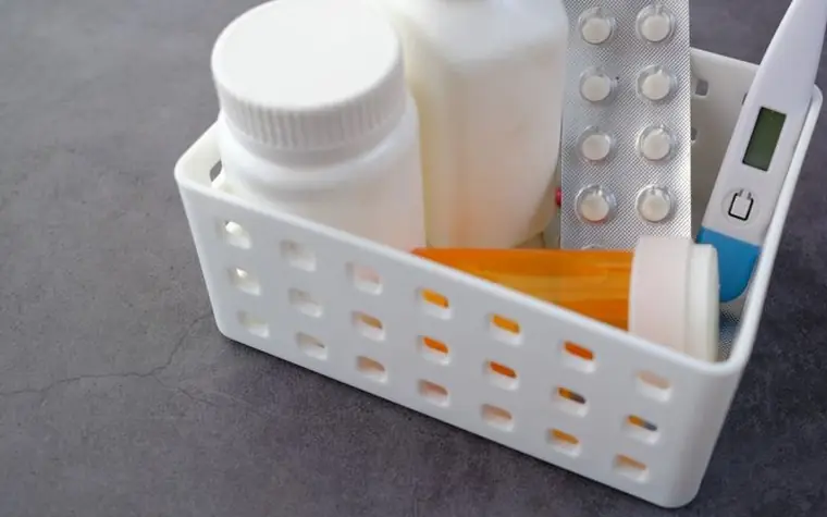 A white basket holding medicine bottles, a blister pack and a thermometer on a dark surface