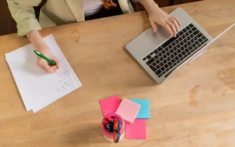 Person writing notes on paper beside a laptop on a wooden desk