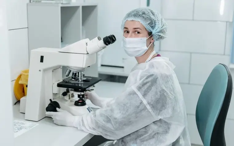 A masked lab researcher looking into a microscope