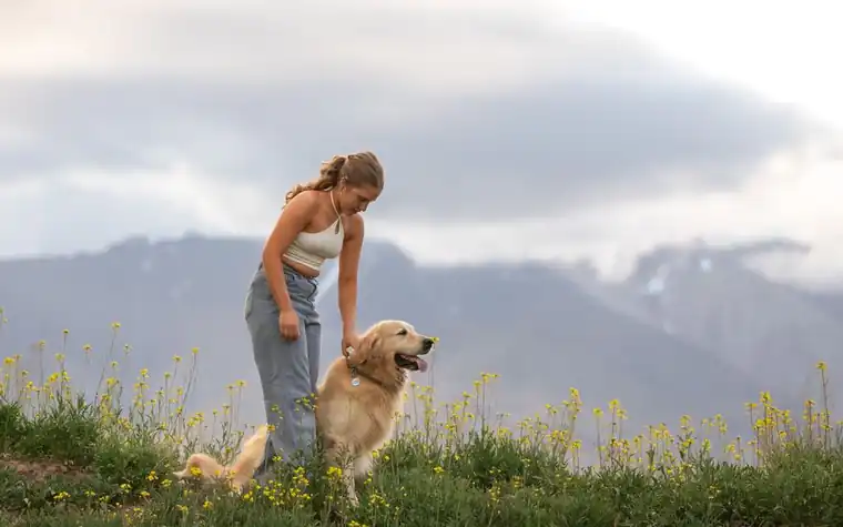 A woman standing in a meadow with a golden retriever beside her and mountains behind