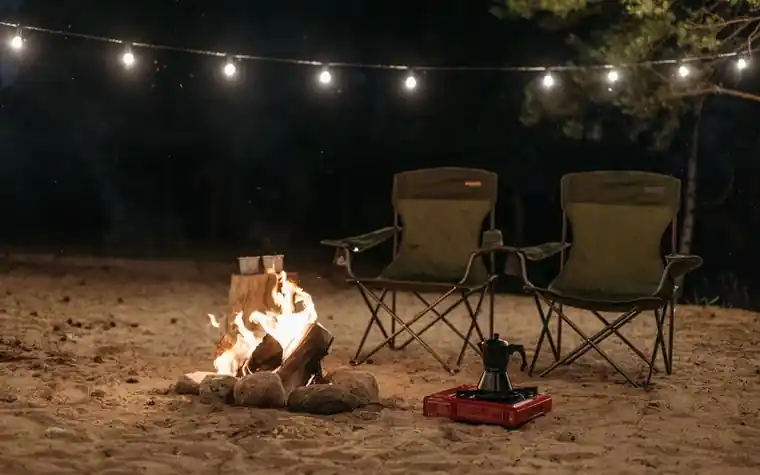 Two camp chairs beside a small campfire under string lights at night