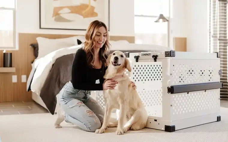 Woman kneeling beside a golden retriever next to an open pet travel crate in a bedroom
