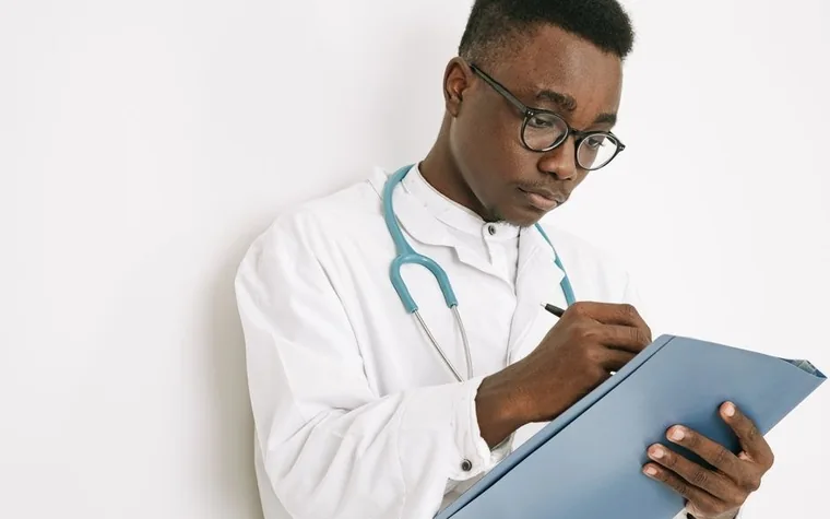 Clinician in a white coat with a stethoscope writing notes on a clipboard