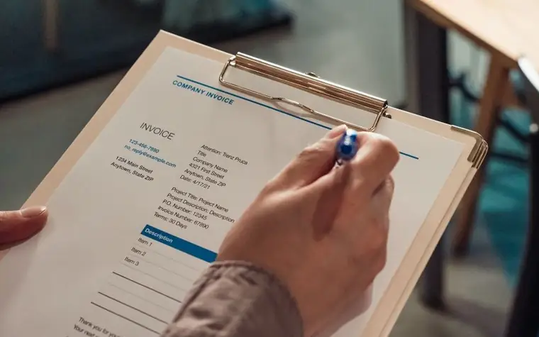 A hand holding a printed invoice attached to a clipboard